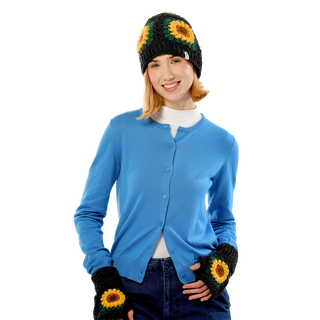 A woman with blonde hair wears a blue button-up cardigan, a white undershirt, and dark jeans. She is accessorized with the Sunflower Sun Hat, which features charming sunflower patterns. With her hands in her pockets and a smile on her face, she stands against a plain background.