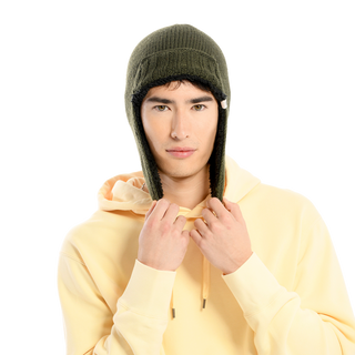 A person wearing a cream-colored hoodie and an Essex Hat in dark green, complete with its extended ear flaps, adjusts it by holding the ear flaps with both hands. The hand-knit materials stand out against the plain black background.