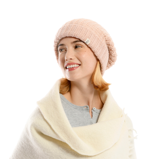 A person with short hair, wearing a pale pink Franklin Hat made of 100% wool and a soft white wrap, looks to the side and smiles. The individual is also dressed in a light gray button-up shirt, perfectly suited for a chic winter wardrobe against the plain black background.