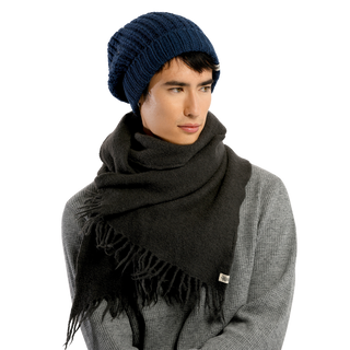 A person is wearing the Franklin Hat, a blue knit beanie, along with a gray sweater and a large dark gray 100% wool scarf. The scarf, which has fringes hanging loosely, is wrapped around their neck and shoulders. They are looking slightly to the side against a plain black background.