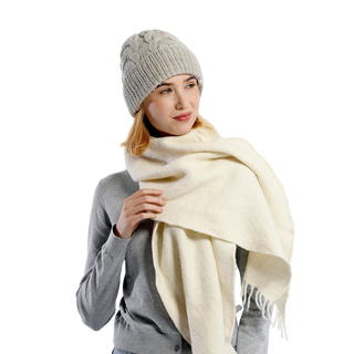 A person wearing a premium Lenox Hat made from gray wool knit, paired with a matching gray sweater and a large cream-colored scarf. The person is holding the scarf with one hand while looking slightly to the side. The plain black background highlights the cozy comfort of their attire.