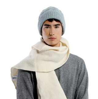 Featuring a neutral expression and an air of cozy comfort, a person stands against a black background, wearing a light blue Lenox Hat paired with a gray sweater and a thick white scarf. They look directly at the camera, combining style with warmth in their outfit.