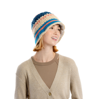 A person wearing a beige cardigan over a light brown top is smiling and looking to the side. They are donning the Stripe Sun Hat, a multicolored, hand-knit bucket hat from Nepal with shades of blue, beige, and red.