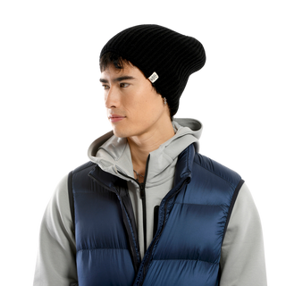 A person looking to their right, wearing a black Tudor Hat made from merino wool, a gray hoodie, and a blue puffed vest. The backdrop is plain.