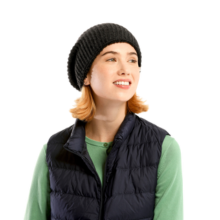 A person with shoulder-length blonde hair is wearing a Tudor Hat, a green long-sleeved shirt, and a black puffer vest. They are looking to the side with a slight smile. The plain background highlights their stylish winter accessory.