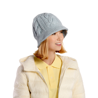 A person with shoulder-length hair is smiling and looking slightly to the side while wearing the Nostrand Hat, a light blue cable knit hat, paired with a yellow collared shirt layered under a light-colored puffer jacket—a charming winter wardrobe. The background is black.