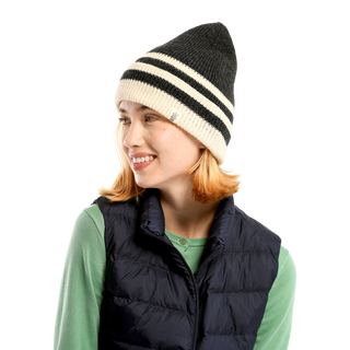 A person with shoulder-length light brown hair wears a black and white McGuinness Hat, a green shirt, and a dark puffer vest. They are smiling and looking to the side against a black background.