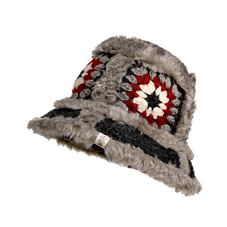 The Crochet Sun Hat With Fur Piping is a round, gray furry hat showcasing patterned crochet panels with red, white, and black designs. It is adorned with faux fur piping and has a small tag peeking out from the base. The background of the image is completely black.