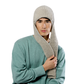 An individual is donning a Pelham Hat, a light gray knitted accessory featuring an elongated, scarf-like extension. Clad in a greenish sweater, they hold part of the merino wool extension in one hand. This beautifully crafted piece was hand-knit in Nepal and stands out against a solid black background.