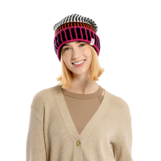 A person with shoulder-length blonde hair is wearing a beige cardigan and an Atlantic Hat, which is a striped multicolor hand-knit Merino Wool beanie. They are smiling and looking directly at the camera against a solid black background.