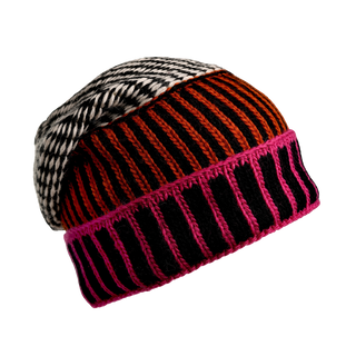 The Atlantic Hat is a multicolored, hand-knit beanie made from Merino Wool that features diverse patterns: a black and white houndstooth design, an orange and black vertical striped pattern, and a section with pink and black horizontal stripes, all set against a black background.