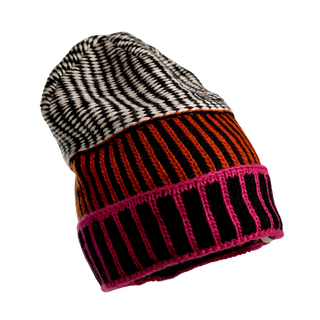 A close-up image of the Atlantic Hat highlights its vibrant, multicolored knitted design. This hand-knit Merino Wool beanie features a top section adorned with black and white diagonal stripes, a middle section displaying red and black vertical stripes, and a bottom section showcasing pink and black vertical stripes.