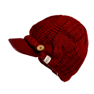 The Broadway Hat is a red cable knit piece featuring a short brim and a decorative wooden button on the side. It includes a small fabric tag with an intricate design, and its thick, interwoven stitches make it a winter essential.