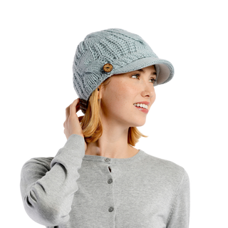 A woman wearing the Broadway Hat, a textured light blue cable knit hat with a small brim and button detail on the side, is looking to her right and smiling. She has light-colored hair and is dressed in a light gray, button-front sweater—a true winter essential ensemble.