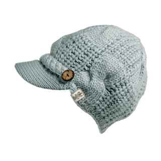 The Broadway Hat is a light blue cable knit cap featuring a visor and a wooden button on the side. It boasts a textured pattern and includes a small fabric tag with an emblem positioned below the button, making it an ideal winter essential.