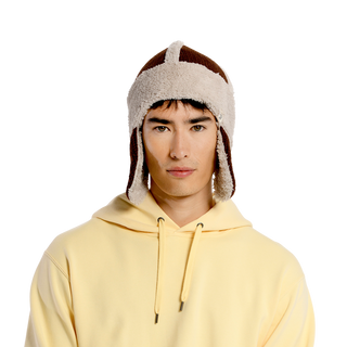 A person wearing a light yellow hooded sweatshirt and the Arthur Hat, a brown and white sherpa-lined aviator winter hat made of cozy Sherpa Fleece. The background is black.