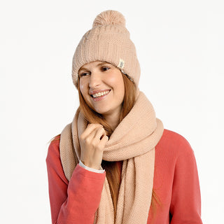 A woman smiles and looks to the side against a white background, wearing the Cedar Hat— a light pink handmade wool beanie with a pom-pom from Nepal—paired with a matching knit scarf over a coral sweater.