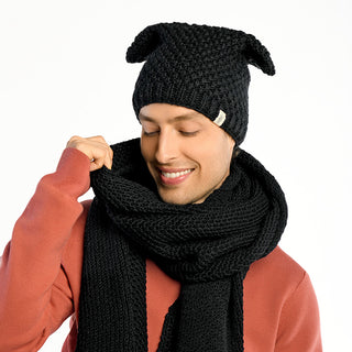 A man smiles with his eyes closed, wearing the Ithaca Scarf handmade in Nepal and a black knit hat with two pointed corners over a salmon-colored sweater, set against a plain white background.
