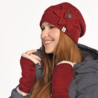 A woman with long brown hair smiles, wearing the Monroe Hat—a red wool beanie handmade in Nepal with a large flower detail—and matching fingerless gloves. She pairs them with a dark puffer jacket and glances slightly to the left.