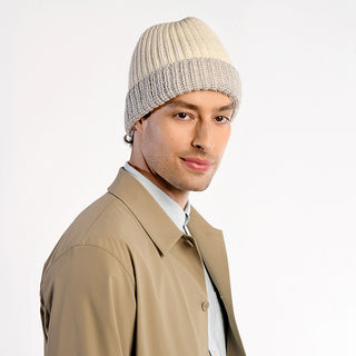 A person wearing the Cliff Hat, a beige and cream wool beanie, along with a light blue shirt and tan trench coat, faces forward and slightly smiles against a plain light background.