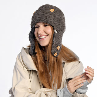 Wearing the handmade Elk Hat—an outdoorsy wool hat from Nepal with ear flaps and wooden buttons—a person with long reddish-brown hair smiles in a light beige jacket and gray top against a plain white background.