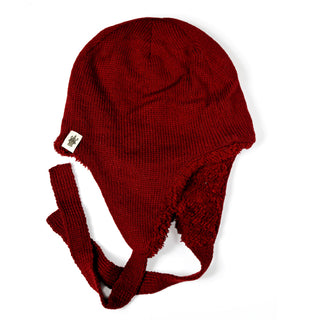 The Hand Knit Snug Hat is a red earflap hat with chin ties, hand knit for warmth and comfort. It features a soft fleece-like lining and a small beige tag on the side. The hat is displayed laid flat on a white background.