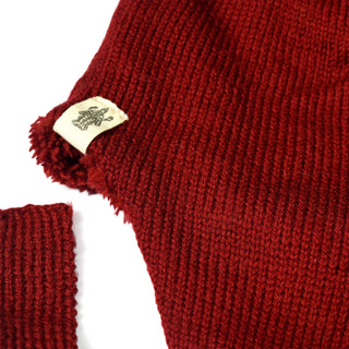 A close-up of red knit fabric from a Hand Knit Snug Hat shows a jagged tear near the edge. A cream-colored tag featuring a small black logo is sewn beside the tear. Another piece of the same cozy winter hat fabric lies nearby.
