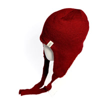 A mannequin head displays the Hand Knit Snug Hat, a deep red earflap hat with two long ties hanging down the sides. Expertly crafted from soft Merino wool, this warm winter accessory features a small fabric tag on the side. The background is white.