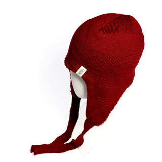 A mannequin head displays the Hand Knit Snug Hat, a deep red earflap hat with two long ties hanging down the sides. Expertly crafted from soft Merino wool, this warm winter accessory features a small fabric tag on the side. The background is white.