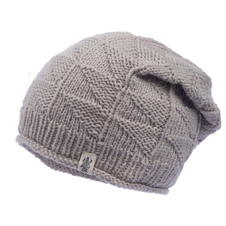 The Hand Knit Wave Slouch, shown in beige merino wool with a geometric pattern, features a folded ribbed edge, small logo tag, and a soft, loosely fitted design.