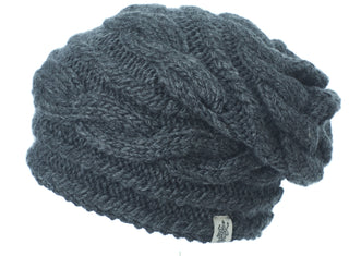 The Triple Braid Cable Slouch is a dark gray, hand-knit Merino Wool beanie from Nepal with a cable pattern, slouchy fit, and a small fabric tag on the brim, shown against a white background.