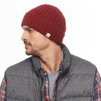 A man with a short beard looks to the side, wearing the Hand Knit Clyde Rib Fold Cap in red, a gray quilted vest, and a plaid shirt over a gray tee against a plain white background.