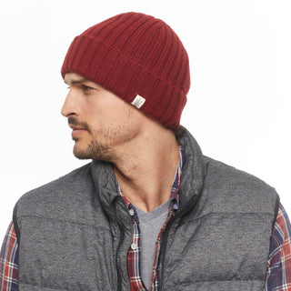 A man with a short beard looks to the side, wearing the Hand Knit Clyde Rib Fold Cap in red, a gray quilted vest, and a plaid shirt over a gray tee against a plain white background.