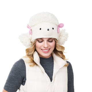 A woman smiles while wearing a whimsical Poodle hat that resembles a sheep, complete with ears and stitched facial features. She has a light vest over a dark shirt.