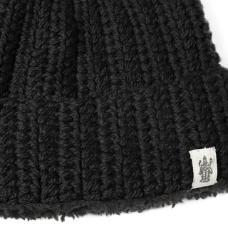 Close-up of the Hand Crochet Chunky Hat, featuring thick, black ribbed knit crafted from lambswool and a folded brim. A small white fabric tag with a printed figure is sewn onto the edge, while the prominently visible texture of the chunky knit highlights its cozy winter cap appeal.