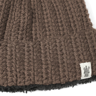 A close-up of the Hand Crochet Chunky Hat, a cozy lambswool beanie with a fold-over brim. A small white fabric tag with a black line drawing of a figure with upraised arms is sewn onto the brim, adding a touch of style to this warm winter accessory.