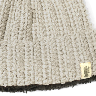 A close-up of the Hand Crochet Chunky Hat, featuring beige chunky knit with a ribbed texture. A small rectangular tag with a black design is sewn onto the edge, and the bottom edge of the beanie is lined with a darker brown yarn for added contrast.