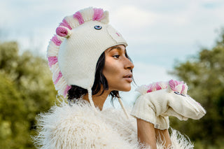 A person wearing a white, hand knit unicorn hat from Nepal with a pink and white mane, matching unicorn mittens, and a fuzzy textured coat.