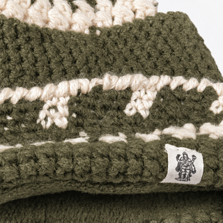 A close-up of the Hand Crochet Discover Hat highlights its patterned olive-green and cream knit with a striped fold-cuff. A white fabric tag featuring a printed logo of two figures and a trophy is sewn onto the brim, adding detail to this soft lambswool accessory.