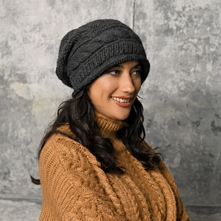 A woman with long dark hair is smiling while looking slightly to the side, wearing a chunky brown knit sweater and the Wave Slouch beanie in dark gray. The background features a textured gray wall.