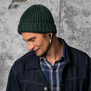 A man wearing the Clyde Rib Fold Cap in green, paired with a plaid shirt and dark denim jacket, stands against a textured gray wall, looking down with a slight smile.