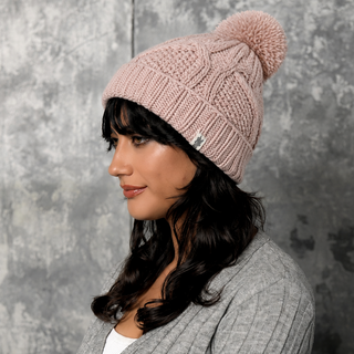 A woman with long dark hair, wearing the Cedar Hat— a handmade in Nepal pink knit wool beanie featuring a pom-pom and faux shearling lining—paired with a grey sweater, stands in front of a textured grey background and looks to the left.