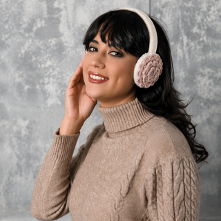 A woman in a beige cable-knit turtleneck sweater wears the Camellia Earmuffs, featuring light-colored faux fur lining, and smiles as she touches one ear with her hand. The background is a soft, gray textured surface.