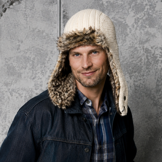 A man with light facial hair and a slight smile stands in front of a gray textured wall, wearing a plaid shirt, denim jacket, knit gloves, and the Winter Trapper Hat—a white winter hat featuring ear flaps and faux fur lining.