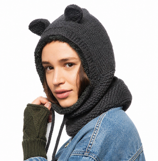 A person wears the Teddy Hood—a dark, handmade merino wool hood from Nepal featuring cozy sherpa lining and small bear-like ears—that covers most of their hair and part of their neck. They also sport a green fingerless glove and a blue denim jacket while looking toward the camera.