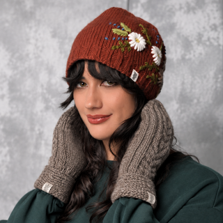 A woman poses in front of a gray backdrop, smiling softly with her hands near her face while wearing cozy Soho Mittens and a rust-colored knit hat adorned with embroidered flowers.