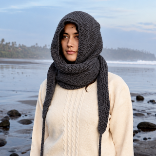 A person wearing a beige sweater and the Hand Knit Anna Hood stands on a rocky beach with waves, palm trees, and a partly cloudy sky in the background.