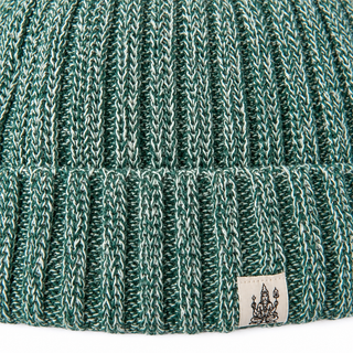 Close-up of the Hand Knit Grid hat, a green and white ribbed knit Merino wool beanie with a folded brim. A small rectangular tag, featuring a seated figure logo and text, is sewn onto the brim.