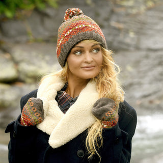 A woman with long blonde hair wears a black coat with a shearling collar and the Hand Knit Explore Hat—a cozy, fleece-lined beanie topped with a pom-pom—along with matching mittens. She stands outdoors in front of rocks, gazing off to the side.