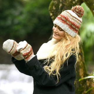 A person with long blonde hair wears the Hand Knit Explore Hat—a Fair Isle beanie with red and green patterns—paired with matching mittens and scarf, along with a dark coat, standing outdoors near a tree and a softly blurred waterfall in the background.